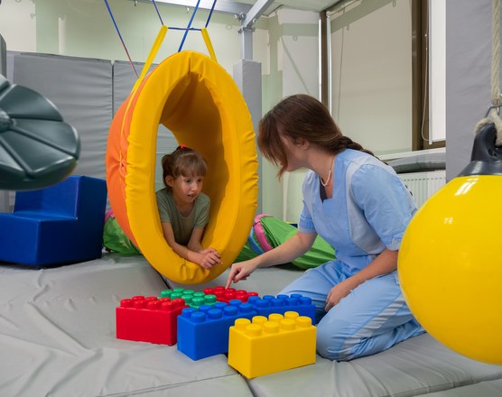 A young child sitting inside a soft play pod while a therapist kneels beside them with building blocks