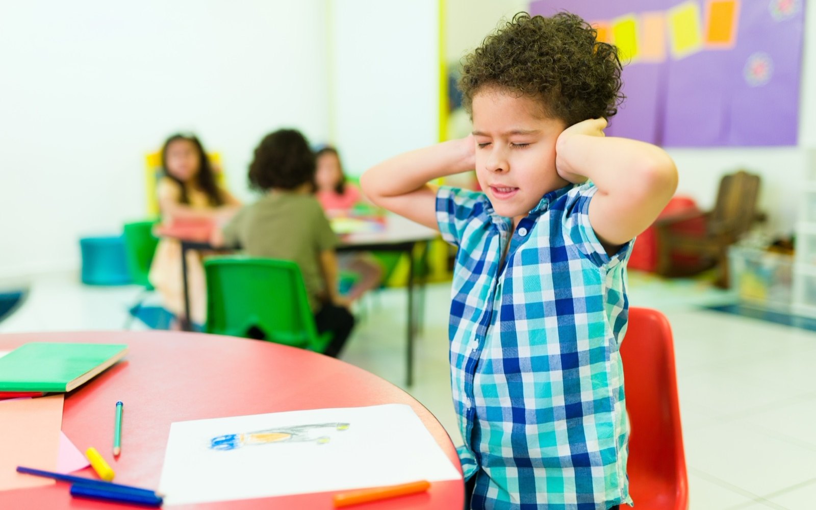 A boy at a classroom table covering his ears with both hands, eyes shut, while other children work behind him