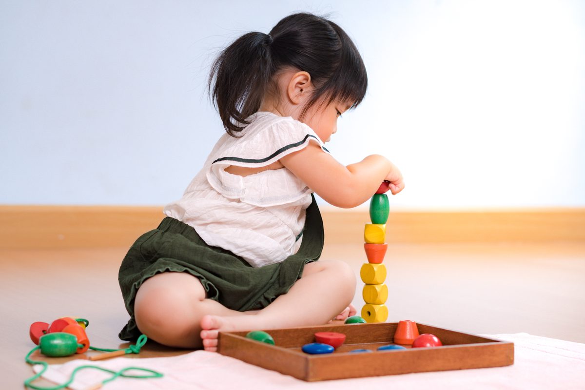 A toddler sitting on the floor stacking large wooden beads into a tower, with a lacing toy beside her