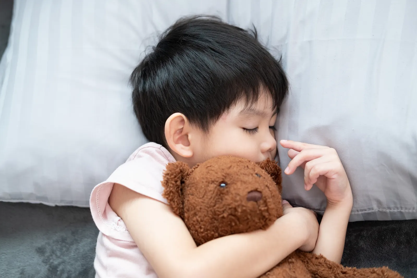 A young child asleep on a striped pillow, holding a teddy bear, breathing through a closed mouth
